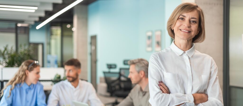 A confident woman in a white blouse stands smiling with arms crossed in the foreground of a modern office, while three colleagues sit and talk in the background.