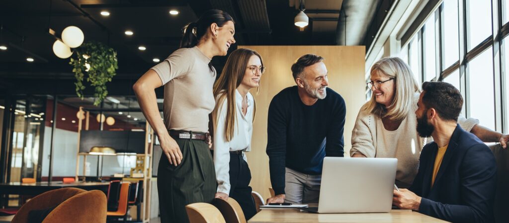 Five colleagues stand and sit around a table in a modern office, smiling and talking as they look at a laptop. Large windows let in natural light and create a friendly, collaborative atmosphere.