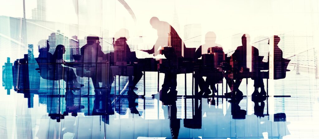 A group of people sit around a conference table in an office, with one person standing and shaking hands. The scene is backlit and reflected on a shiny surface, creating a double exposure effect.