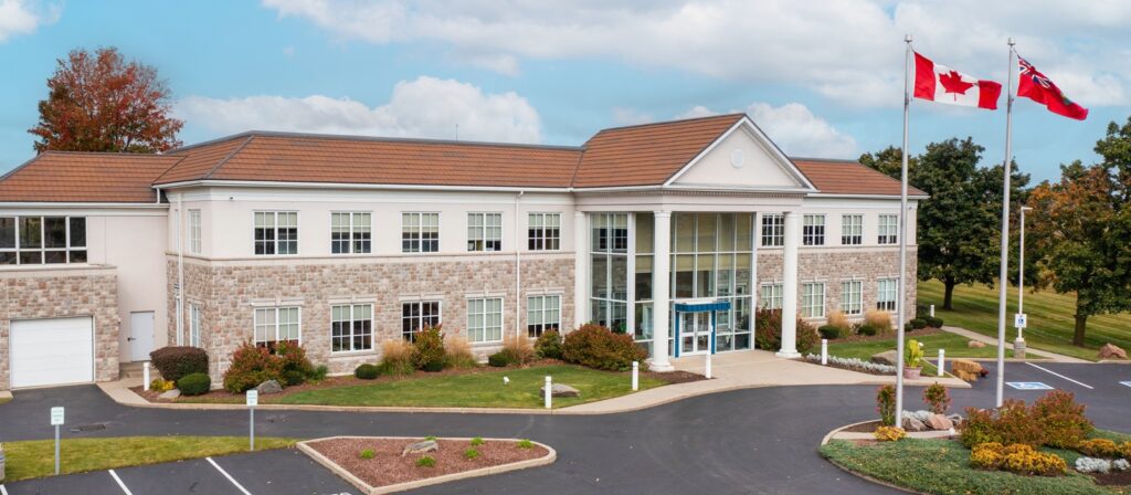 A two-story stone and white building with large windows and columns at the entrance, two Canadian flags flying outside, surrounded by trees, shrubs, and a parking lot under a partly cloudy sky.