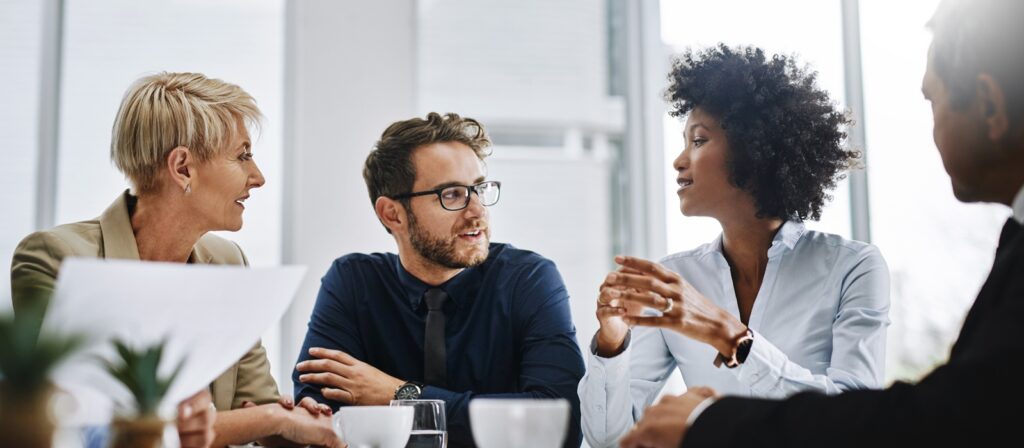 Four professionals sit at a conference table in an office, engaged in discussion. Two women and two men listen intently, with documents and coffee cups in front of them. Bright natural light fills the room.