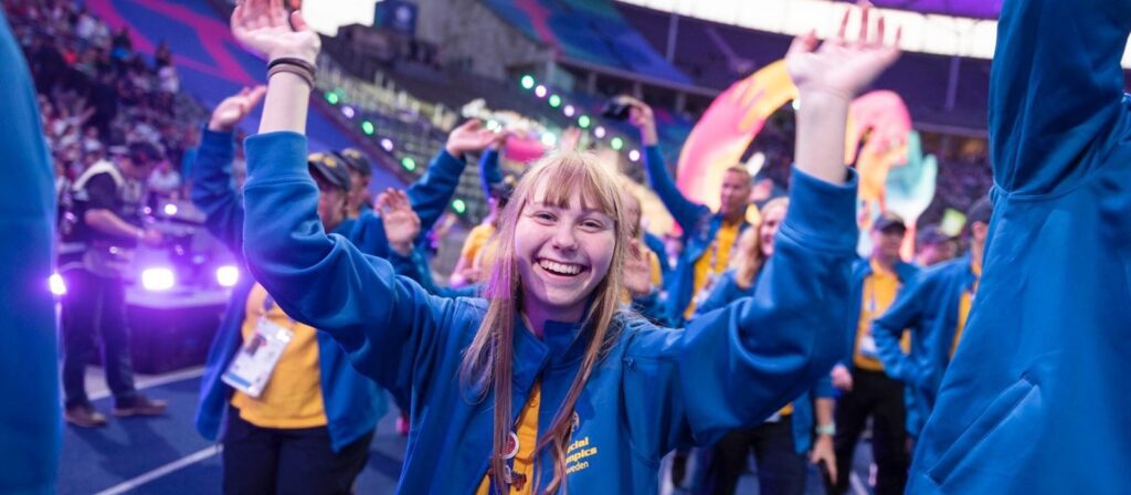A young woman in a blue hoodie smiles and raises her arms in celebration at a lively stadium event, surrounded by other people in similar outfits and colorful lights.