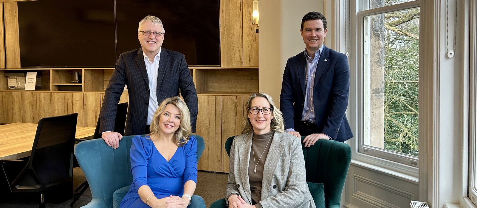 Four professionally dressed people, two men standing and two women sitting in armchairs, pose and smile inside a modern office with large windows and wooden decor.