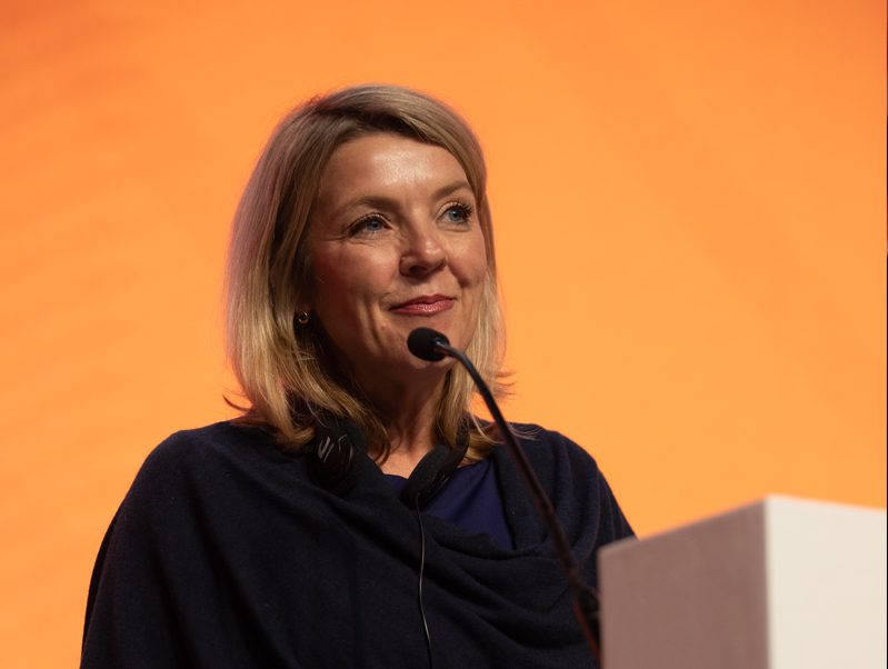 A woman with blonde hair speaks into a microphone at a podium, smiling slightly. She wears a dark top and the background is a solid orange color.
