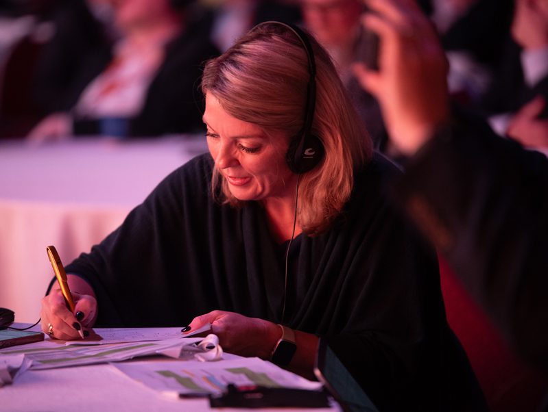 A woman wearing headphones sits at a table, smiling and writing notes with a pen on paper. She appears focused, with people blurred in the background of a conference or meeting setting.