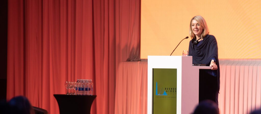 A woman stands at a podium, speaking into a microphone at an event. Behind her is a large orange screen and pink curtains. Bottled water sits on a table nearby. The podium displays a Buenos Aires Americas sign.