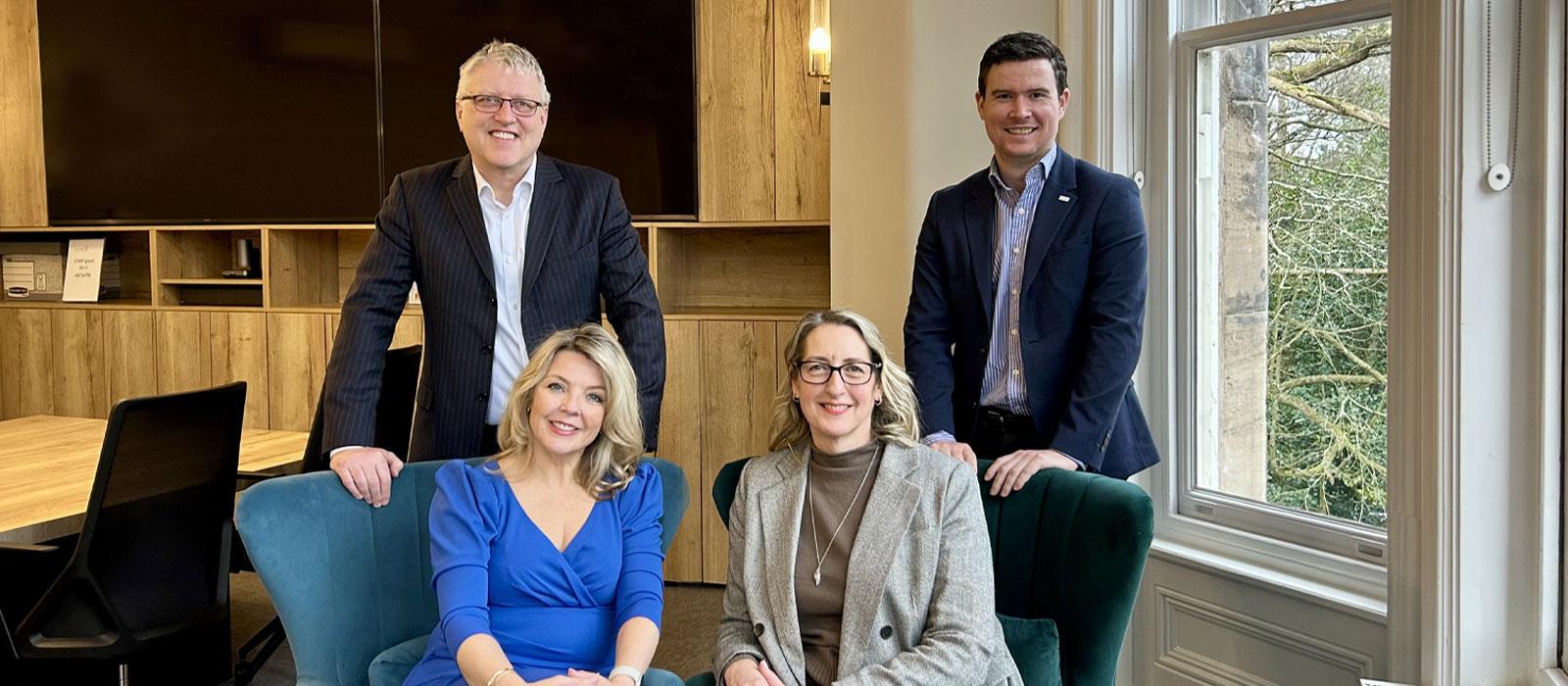 Four professionally dressed adults, two standing and two seated, pose and smile in a modern office with wood-panel walls and large windows showing trees outside.