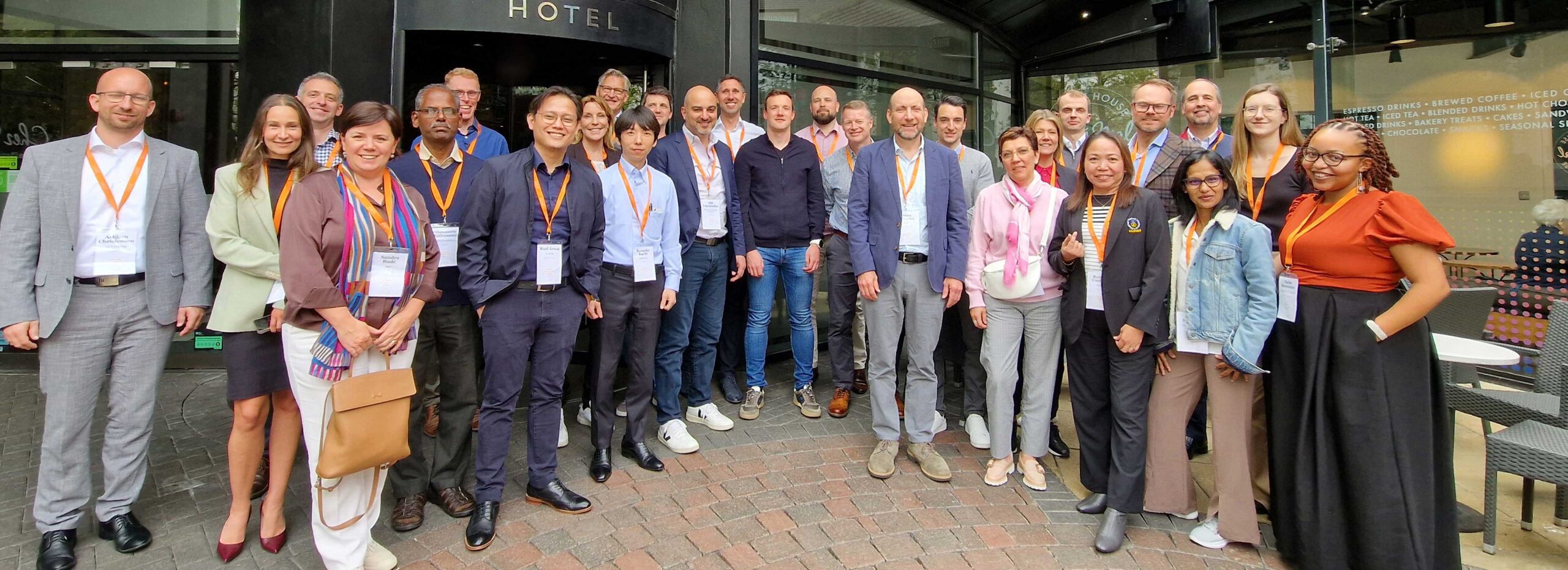 A diverse group of around 25 people, many wearing conference lanyards, pose and smile together outside a hotel entrance on a brick pavement.