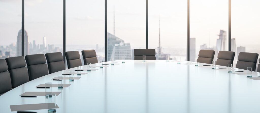 Modern conference room with a long glass table, black chairs, notepads, and glasses, set in front of large windows overlooking a city skyline with tall buildings.