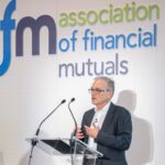 A man in a suit speaks at a podium with microphones in front of a backdrop that reads association of financial mutuals.