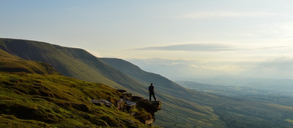 A person stands on a rocky ledge overlooking rolling green hills and a sunlit valley under a partly cloudy sky.