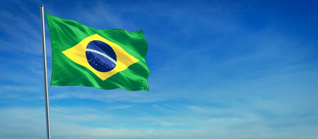 The flag of Brazil waving on a flagpole against a bright blue sky with light clouds. The flag is green with a yellow diamond and a blue globe featuring stars and a white banner.