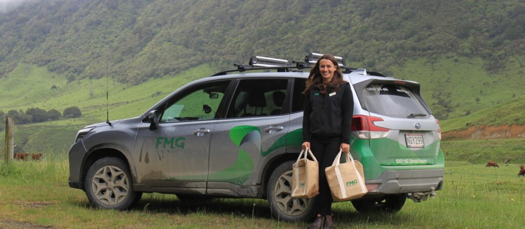 A woman stands next to a green and silver FMG-branded SUV parked on a grassy field with hills in the background, holding two FMG tote bags and smiling at the camera.