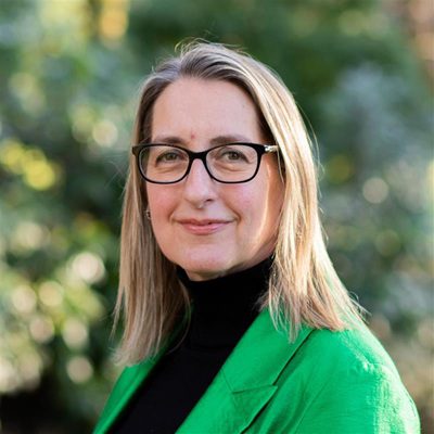 A woman with straight blonde hair and glasses, wearing a black turtleneck and bright green blazer, smiles outdoors with blurred greenery in the background.