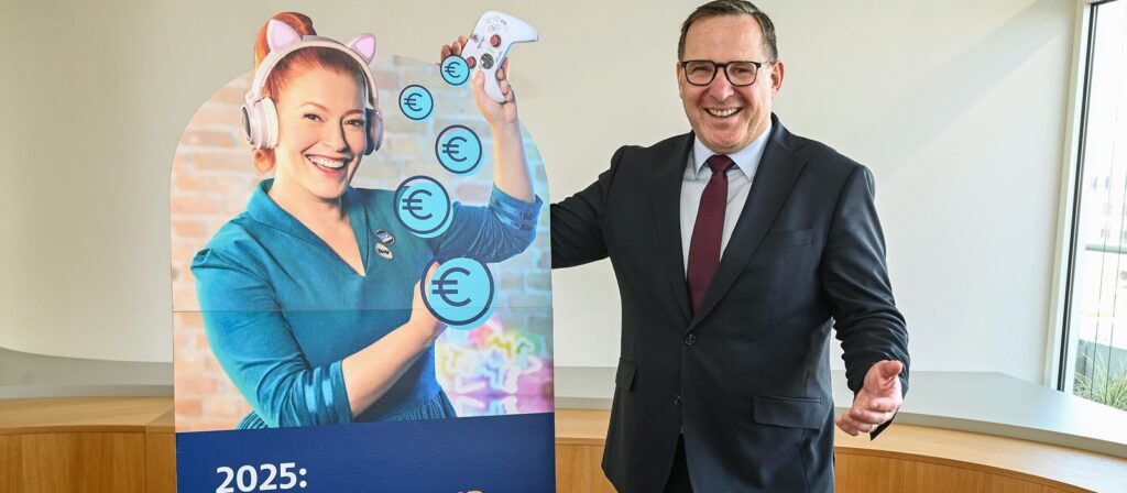 A smiling man in a suit stands beside a cardboard cutout of a cheerful woman wearing cat ear headphones, holding a game controller with euro symbols. The cutout displays 2025 and text below.