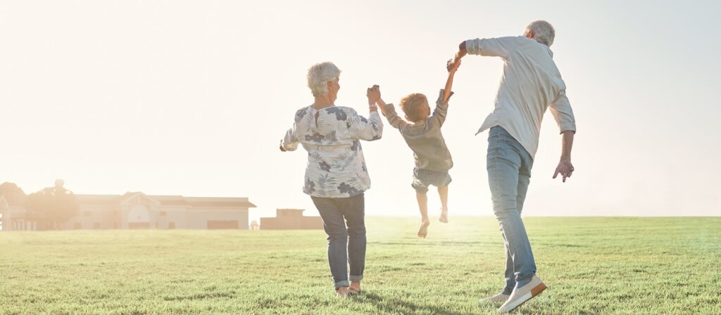 Two older adults swing a young child by the arms as they walk on a grassy field in the sunlight, with houses visible in the background.