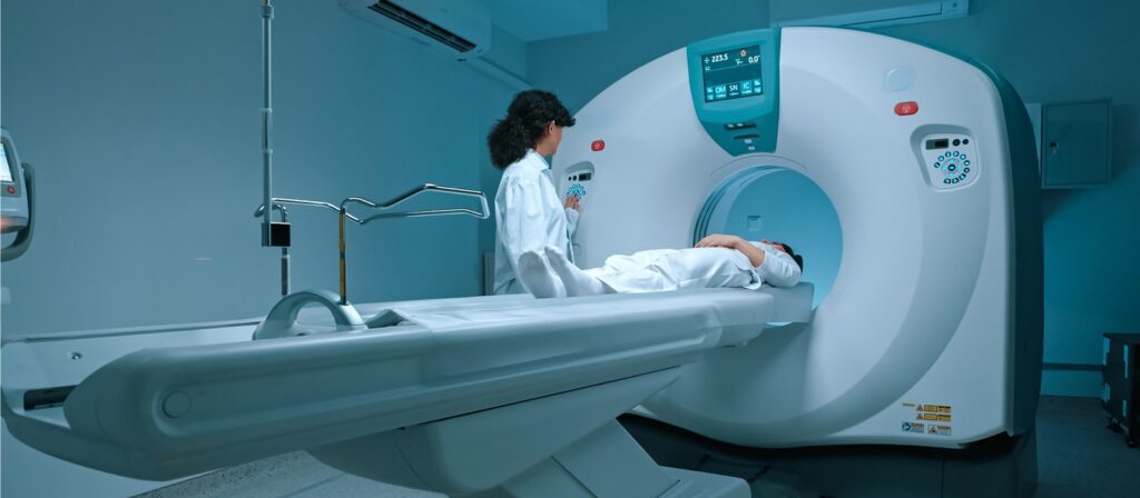 A medical technician stands beside a patient who is lying on a CT scan machine bed, preparing for a scan in a modern, sterile hospital room with blue lighting.