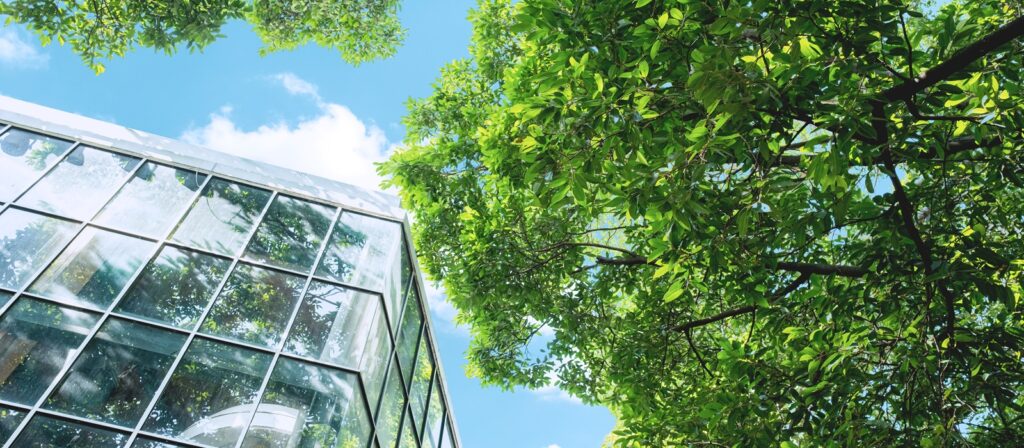 Glass building façade reflects lush green trees under a bright blue sky with clouds, emphasising a blend of nature and modern architecture.