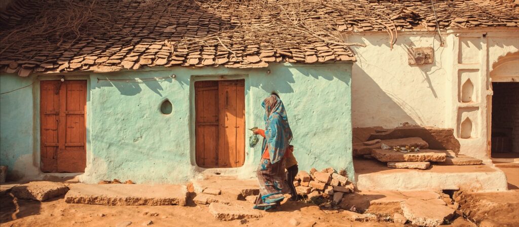 A woman in traditional clothing walks past an old, rustic house with light blue walls, wooden doors, and a tiled roof. Sunlight casts warm shadows on the building and uneven ground.