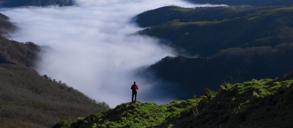 A person in a red jacket stands on a grassy hillside overlooking a valley filled with white clouds, surrounded by dark, tree-covered mountains under a clear sky.