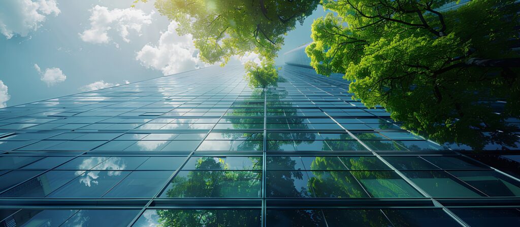 A low-angle view of a modern glass skyscraper reflecting the blue sky, white clouds, and green trees, with sunlight shining through the tree branches.
