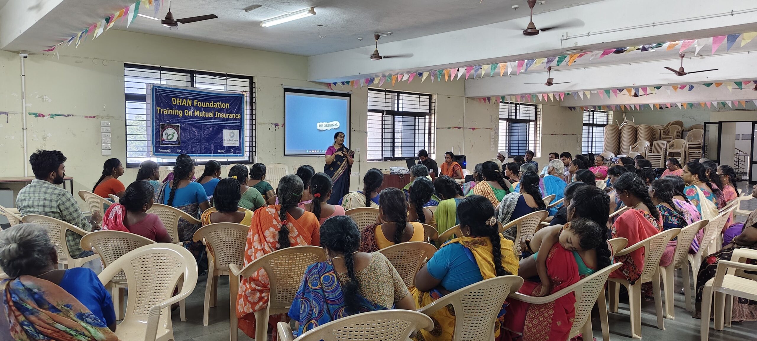A woman stands and speaks to a large group of people seated in plastic chairs at an indoor training session. A banner reads DHAN Foundation Training on Mutual Insurance. A projector screen shows a presentation.