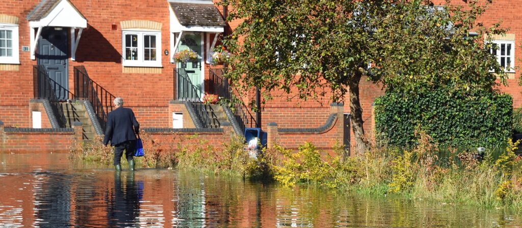 A person wearing wellies walks through floodwater in front of two brick houses with steps leading up to their doors. The water covers the road and some vegetation, and a tree stands nearby.
