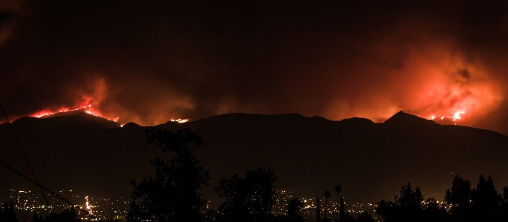 Wildfires burn along a mountain ridge at night, emitting bright orange flames and smoke, while city lights shine below in the foreground.