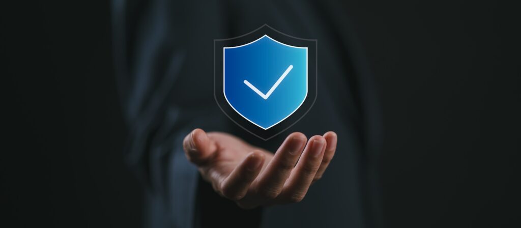 A person extends their open hand forward with a digital blue shield icon and a tick hovering above the palm, symbolising security and protection. The background is dark and blurred.