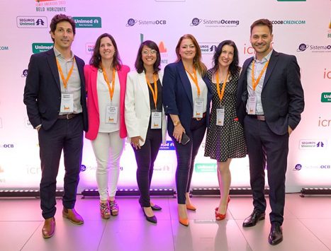 Six professionally dressed people, wearing name badges and orange lanyards, stand smiling together in front of a white event backdrop with multiple logos.