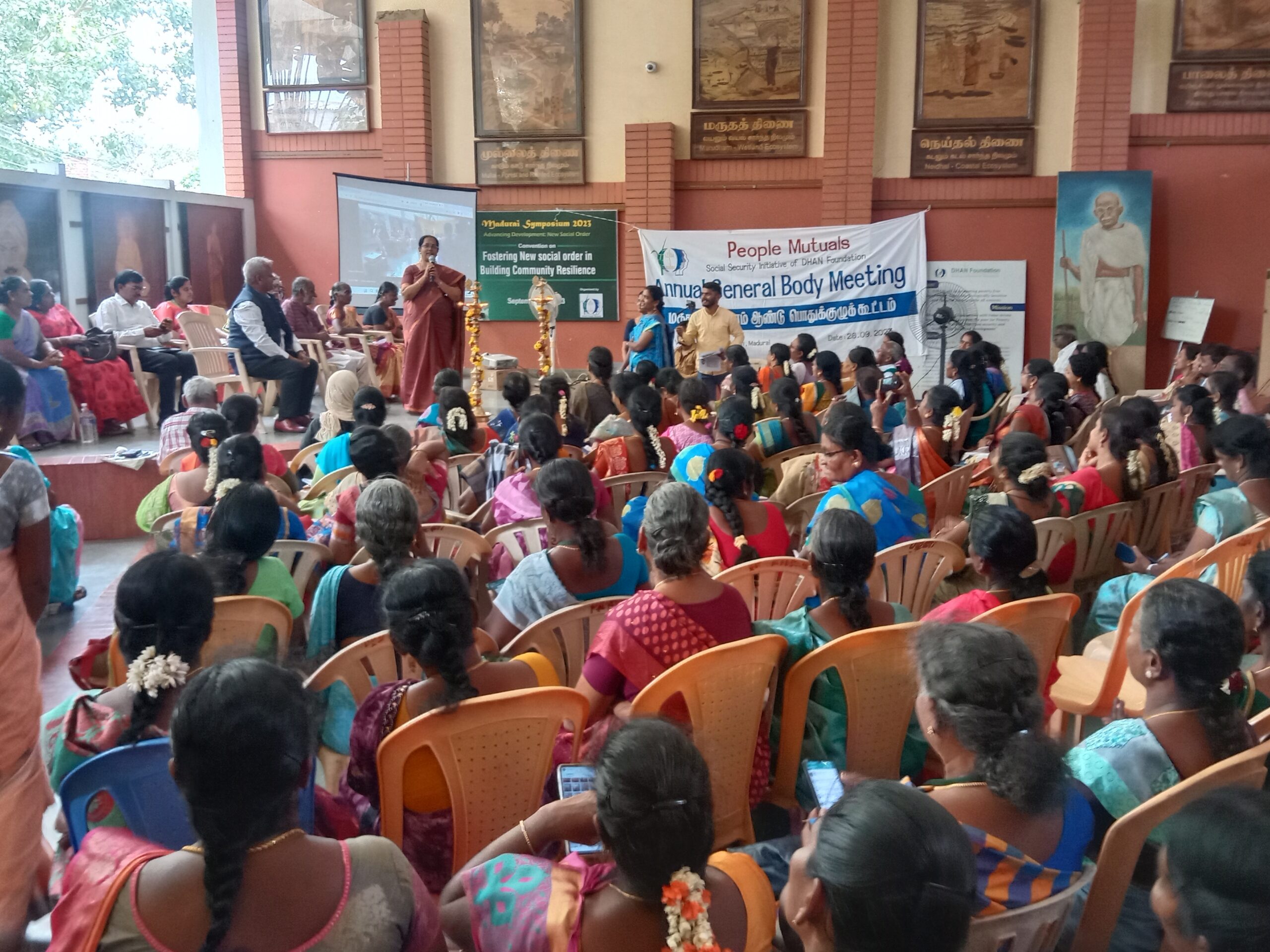 A large group of women sit on plastic chairs, facing a stage where speakers address them at a community meeting. A banner reads Annual General Body Meeting. The event appears to be held indoors, with wall art decor in the background.