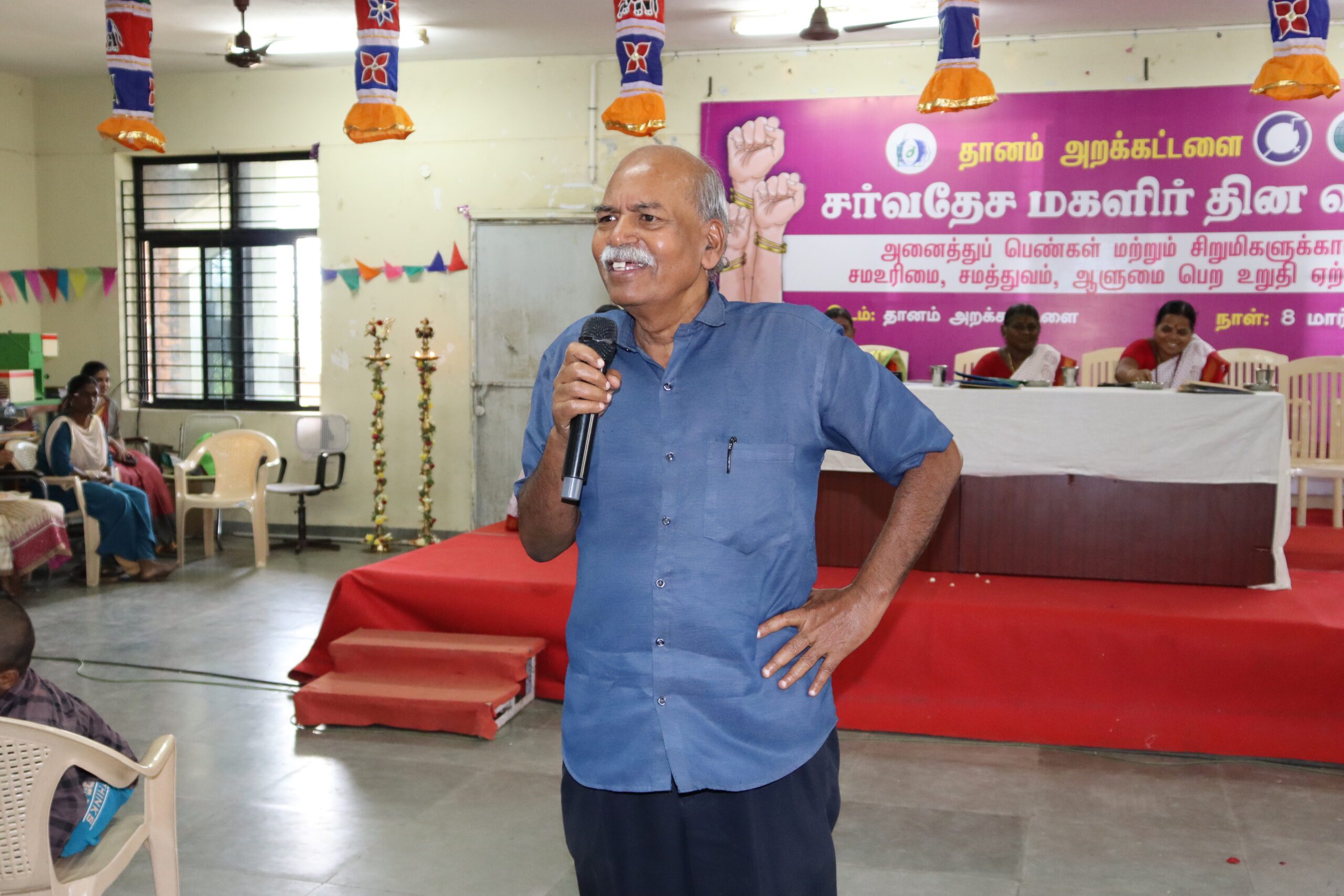 An older man with grey hair and a moustache, wearing a blue shirt, speaks into a microphone in a decorated hall with people seated and a stage in the background featuring a pink banner in Tamil.