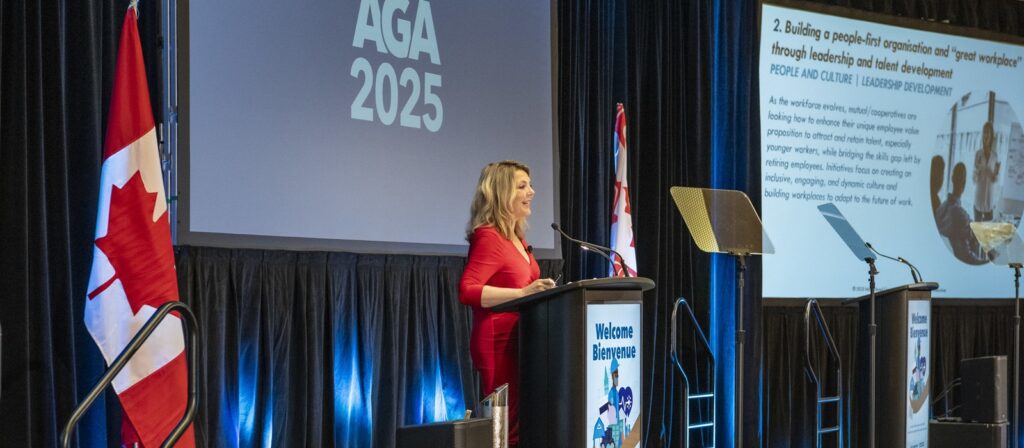 A woman in a red dress speaks at a podium on a stage with Canadian flags, large screens displaying “AGA 2025,” and a presentation slide about leadership and workplace development.