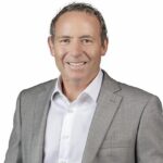 A middle-aged man with short, curly hair smiles at the camera. He is wearing a light grey suit jacket over a white dress shirt. The background is plain white.