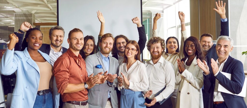 A diverse group of professionals smiles, cheers, and claps together in an office setting, celebrating in front of a projection screen, conveying teamwork and success.