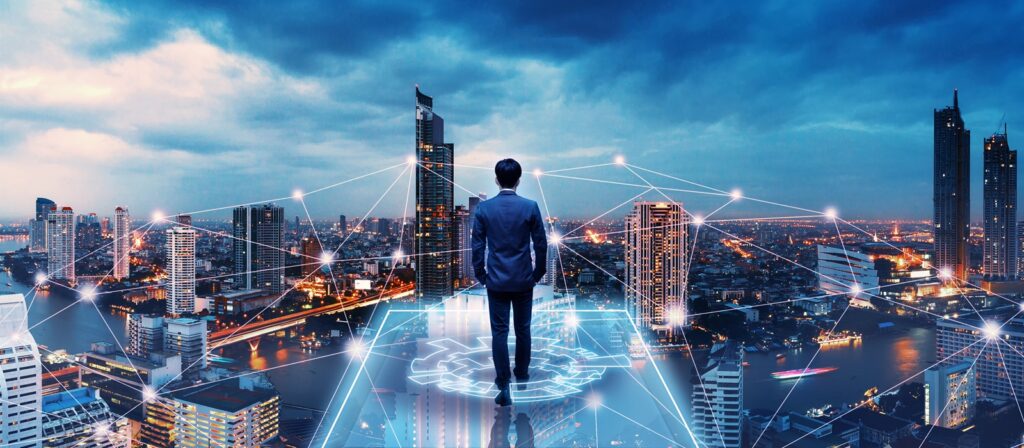 A man in a suit stands on a digital platform overlooking a cityscape at dusk, with connected lines and digital elements overlaying the buildings, symbolising technology and connectivity.