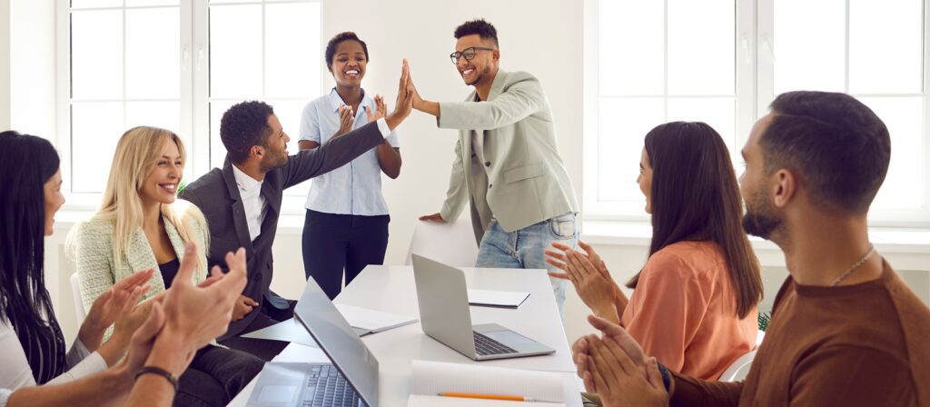 A group of six diverse colleagues sit around a table, smiling and clapping as two people in the centre give each other a high five in a bright office setting. Laptops and papers are on the table.