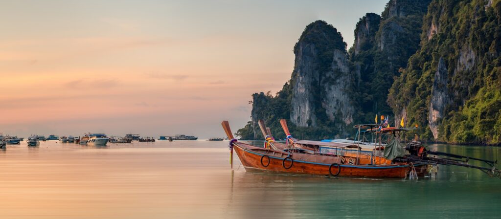 A traditional wooden longtail boat floats on calm water at sunset, with several other boats in the distance and steep, tree-covered limestone cliffs in the background. The sky is a soft gradient of pink and orange.