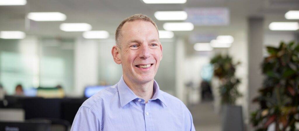 A man in a light blue shirt smiles whilst standing in a bright, modern office with blurred desks, people, and plants in the background.