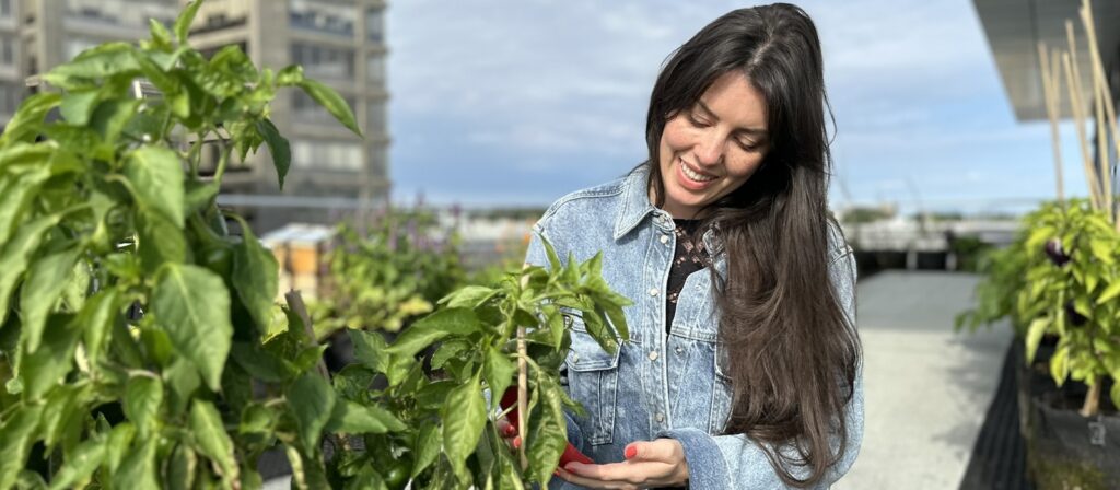 A woman with long brown hair, wearing a denim jacket, smiles as she tends to green plants on a rooftop garden with buildings and blue sky in the background.