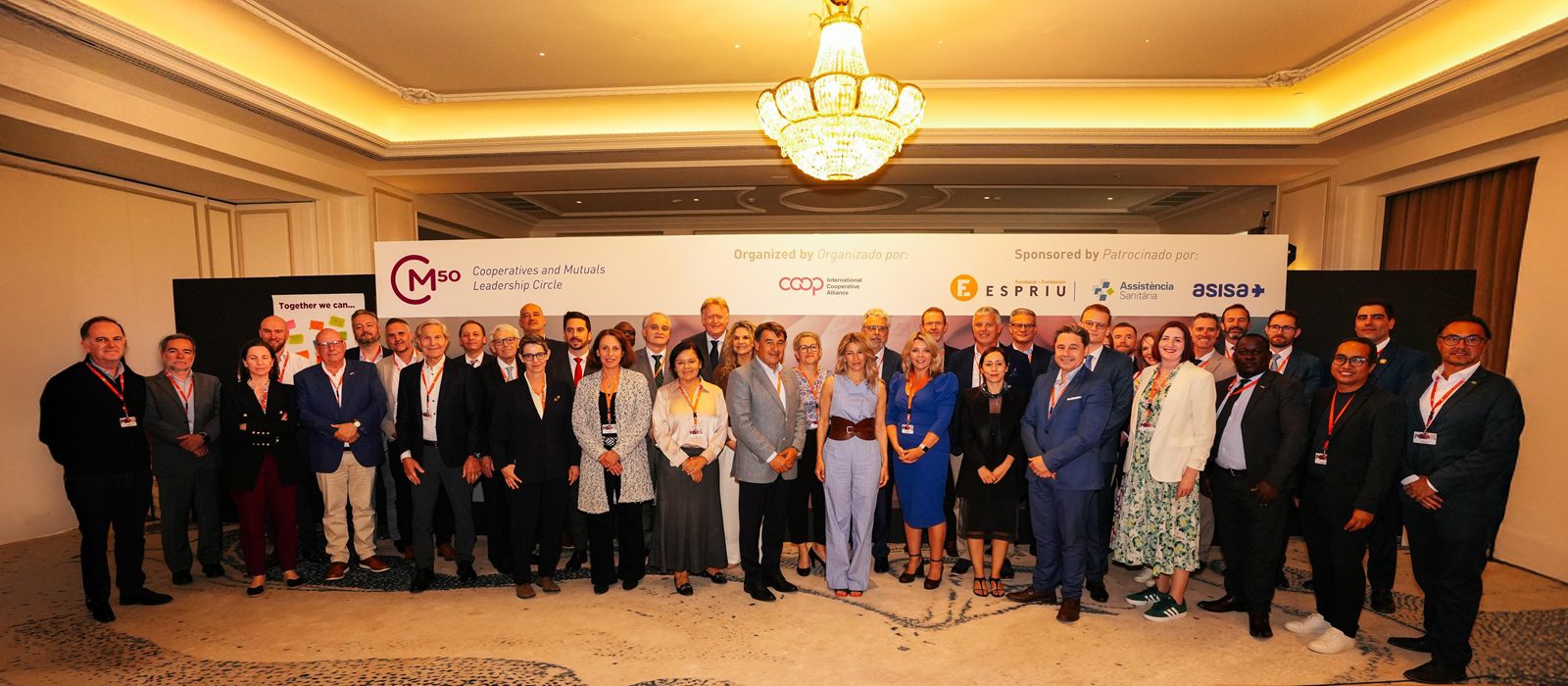 A large group of professionally dressed people stand together in a well-lit conference room, posing for a group photo in front of banners with various organisational and sponsor logos.