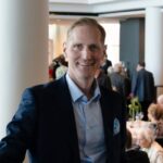 A man in a navy suit and light blue shirt smiles at the camera in a bright, modern room with large windows and people socialising in the background.