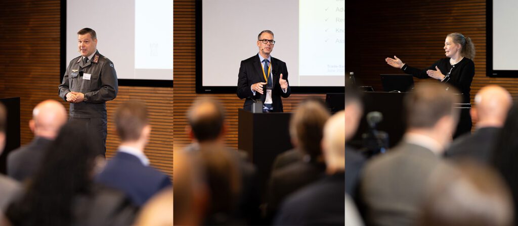 Three speakers address an audience at a conference: a uniformed officer, a man in a suit, and a woman gesturing at a lectern, with a wooden wall and presentation screen in the background.