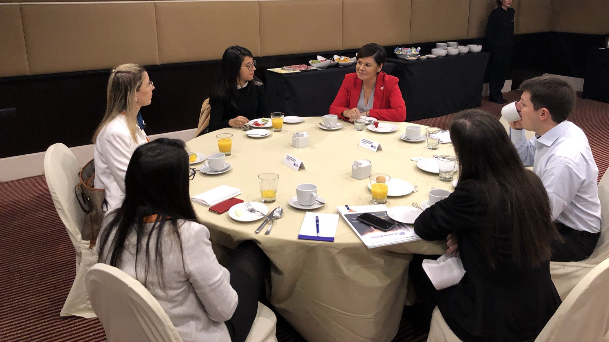 Six people sit around a round table covered with a beige tablecloth, having a discussion. There are drinks, notepads, and breakfast items on the table. A buffet with dishes is visible in the background.