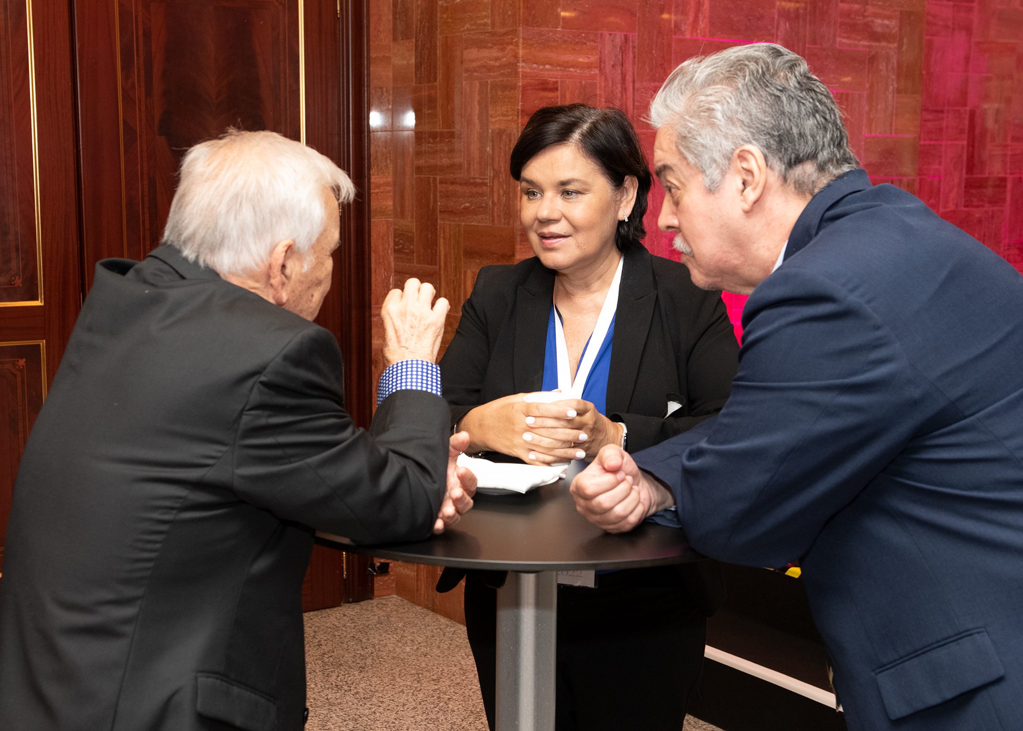 Three people in business attire are standing around a small round table, engaged in a serious conversation. The woman in the middle is holding a cup, whilst the two men lean in and listen attentively.