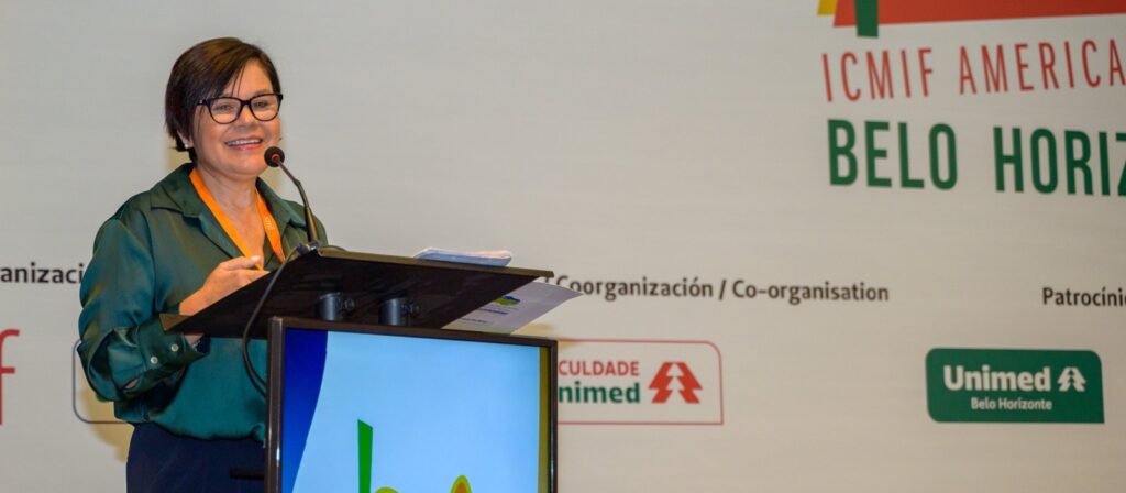 A woman wearing glasses and a green blouse speaks at a lectern with a microphone during the ICMIF Americas event in Belo Horizonte. The background displays event information and sponsor logos.