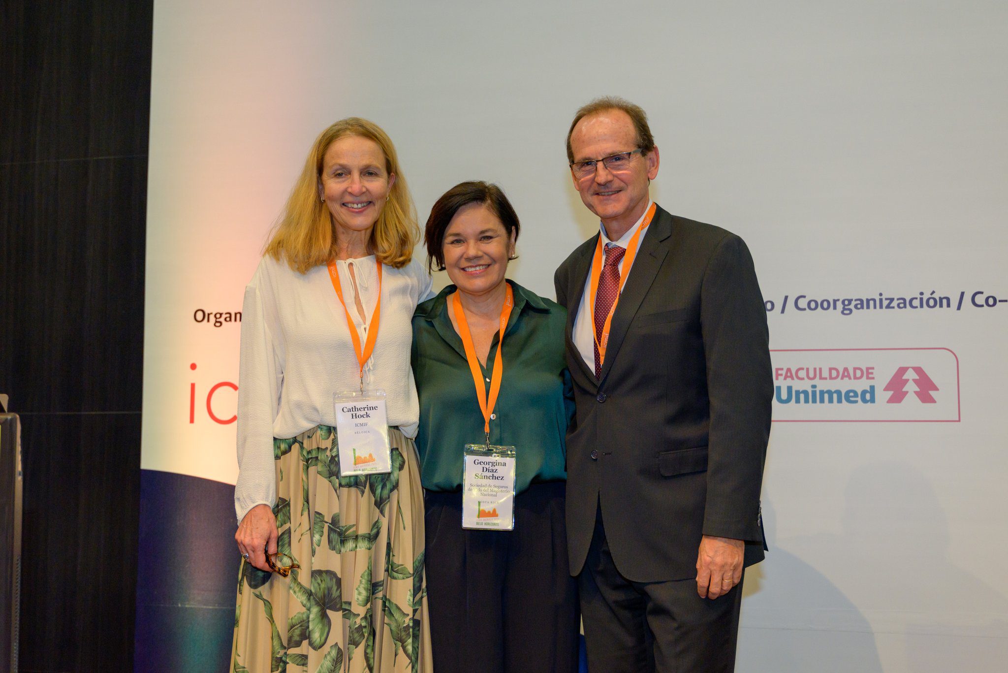 Three people stand together indoors, smiling at the camera and wearing conference badges with orange lanyards. The background features a white banner with logos and text.