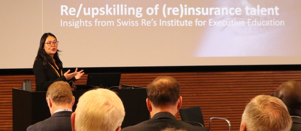 A woman stands at a lectern giving a presentation to an audience. The screen behind her displays the title, Re/upskilling of (re)insurance talent: Insights from Swiss Re’s Institute for Executive Education.