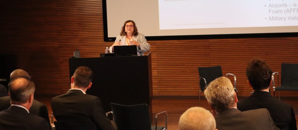 A woman stands at a lectern giving a presentation to an audience in a conference room. A projection screen with bullet points is visible behind her, and attendees sit facing the stage.