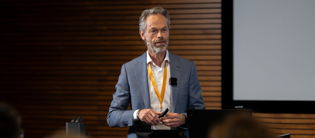 A man with grey hair and a beard, wearing a blue suit and orange lanyard, stands and speaks in front of an audience in a conference room with wooden panelling and a projection screen.